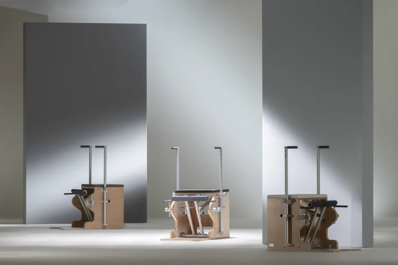 Drei Pilates-Chairs von BASI Systems mit gepolsterter Sitzfläche, Pedal und zwei Haltegriffen, vor hellen und grauen Studiowänden.